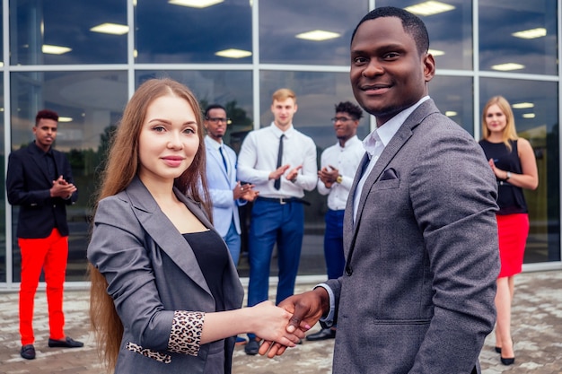 Concept de bonne affaire. Groupe multinational de jeunes gens d'affaires salutations et poignée de main avec des collègues en arrière-plan. Belle femme d'affaires souriante avec un partenaire afro-américain se serrant la main