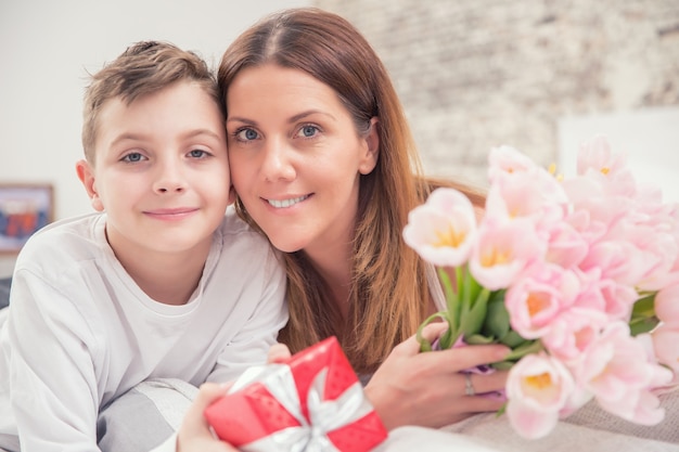 Concept de bonne fête des mères. Maman avec fils sur lit avec cadeau et tulipes.