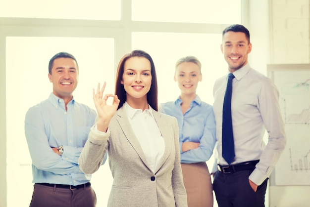 concept de bureau, d'entreprise et de travail d'équipe - sympathique jeune femme d'affaires souriante avec une équipe à l'arrière montrant le signe ok