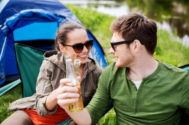 concept de camping, de voyage, de tourisme, de randonnée et de personnes - amis heureux faisant tinter des bouteilles de verre et buvant du cidre ou de la bière au camping