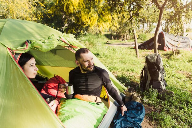 Concept de camping, de voyage, de tourisme, de randonnée et de personnes - couple calme de touristes buvant du café dans des tasses dans la tente et regardant à l'extérieur. Heureux voyageurs en sac de couchage.