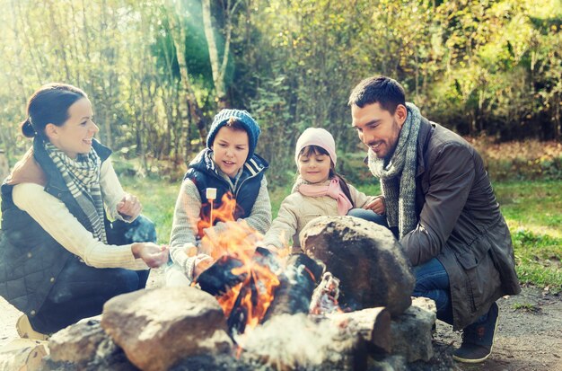 concept de camping, de voyage, de tourisme, de randonnée et de personnes - famille heureuse rôtissant de la guimauve au feu de camp