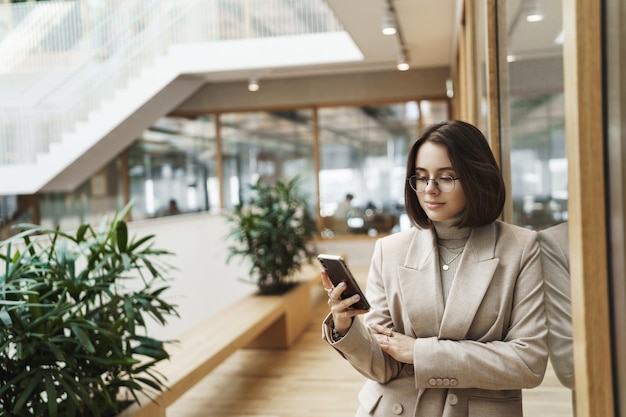 Concept de carrière de femmes de style de vie Goodlooking jeune femme entrepreneur femme de bureau en veste élégante attendant une personne dans le hall de l'hôtel vérifiant les messages sur smartphone souriant au téléphone