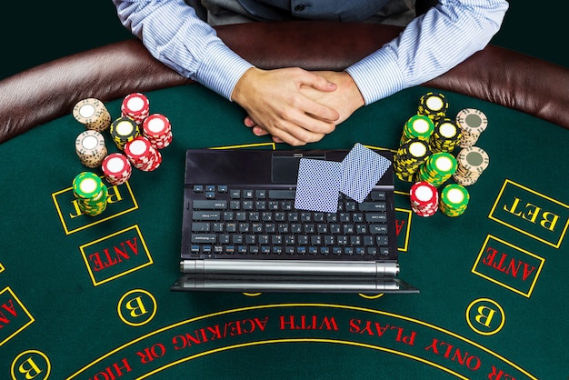 Concept de casino, de jeu en ligne, de technologie et de personnes - gros plan sur un joueur de poker avec des cartes à jouer, un ordinateur portable et des jetons à la table de casino verte