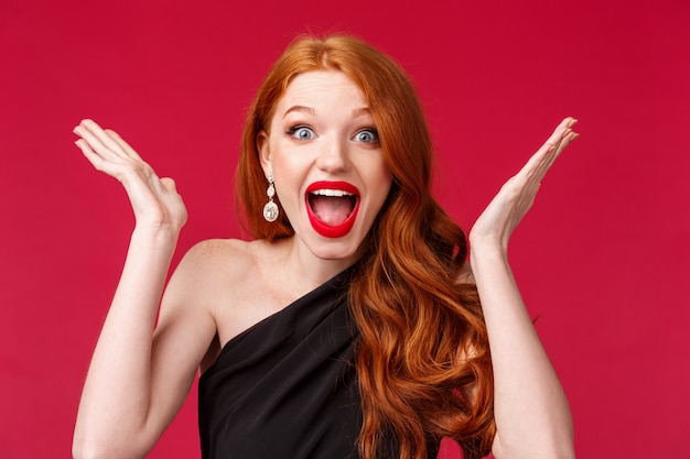 Concept de célébration, d'émotions et de beauté. Close-up portrait of happy cheerful redhead woman winner, triomphant d'excellentes bonnes nouvelles, levez la main et criez joyeux, sur un mur rouge