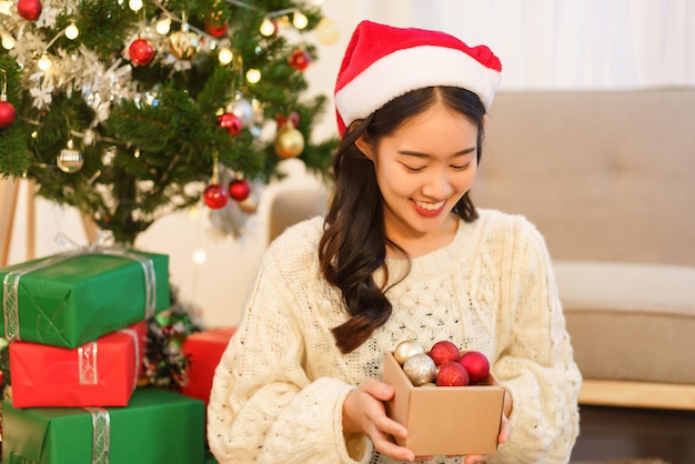 Concept de célébration de noël Une femme asiatique en bonnet de noel tient une boîte de boule de noël pour la décoration