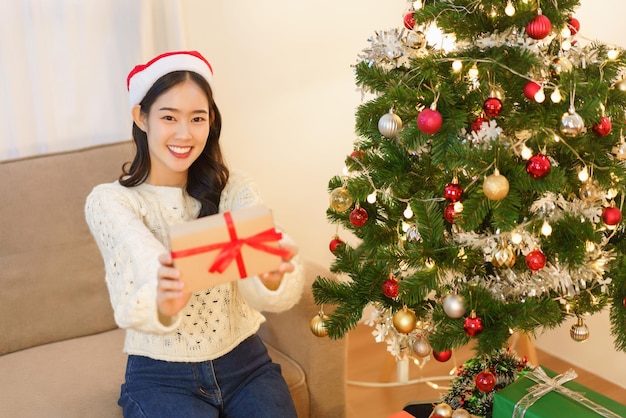 Concept de célébration de Noël Jeune femme asiatique en bonnet de Noel souriant et tenant un cadeau de Noël