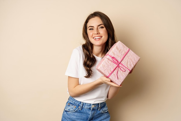 Concept de célébration et de vacances. Heureuse jeune femme tenant un cadeau emballé dans une boîte rose, recevoir un cadeau, l'air étonné et surpris, debout sur fond beige.
