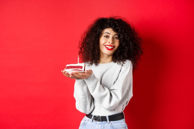 Concept de célébration et de vacances. Sourire belle femme célébrant son anniversaire, tenant le gâteau b-day avec bougie et faire des vœux, debout heureux sur le mur rouge.