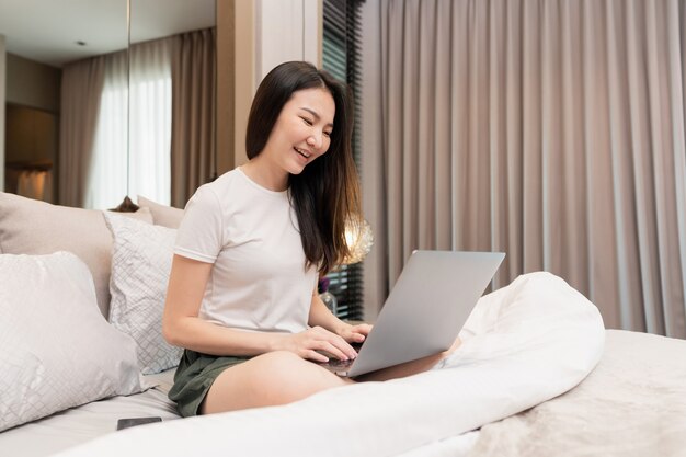 Concept de chambre à coucher dans la chambre à coucher avec l'atmosphère chaleureuse d'une fille assise sur le lit avec la grande couverture utilisant l'ordinateur portable pour travailler en ligne.