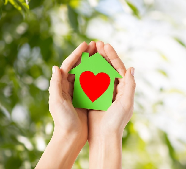 concept de charité, d'immobilier et de maison familiale - photo en gros plan de mains féminines tenant une maison en papier vert avec un coeur rouge