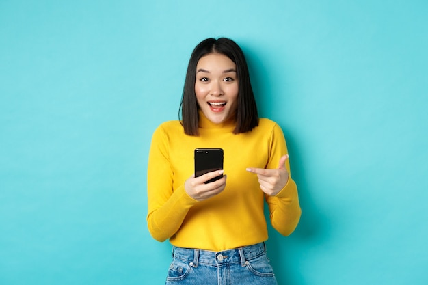 Concept de commerce électronique et d'achat en ligne. Une femme asiatique surprise démontre une application pour smartphone, des remises sur Internet, un doigt pointé sur un téléphone portable, un fond bleu.