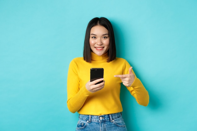 Concept de commerce électronique et d'achat en ligne. Jolie femme asiatique en pull jaune pointant sur le smartphone, souriant à la caméra, debout sur fond bleu.