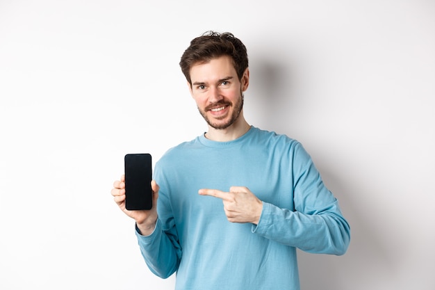 Concept de commerce électronique et de magasinage. Homme caucasien souriant, pointant le doigt sur un écran de smartphone vide, montrant une offre en ligne, fond blanc.