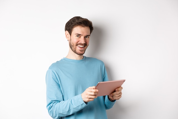 Concept de commerce électronique et de technologie. Jeune homme caucasien souriant à la caméra, tenant une tablette numérique, debout sur fond blanc