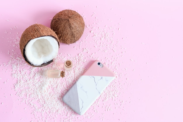 Concept de cosmétiques biologiques à la noix de coco sur fond rose. Noix de coco fraîche, huile de coco et smartfon moderne dans un étui élégant, vue de dessus.