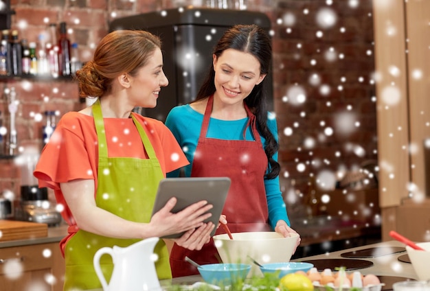 concept de cours de cuisine, d'amitié, de nourriture, de technologie et de personnes - femmes heureuses avec un ordinateur tablette dans la cuisine sur l'effet de neige