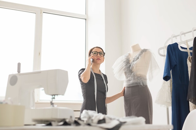Concept de couturière, tailleur, mode et salle d'exposition - une belle jeune créatrice parle au téléphone portable et sourit tout en examinant son travail dans un studio de couture.