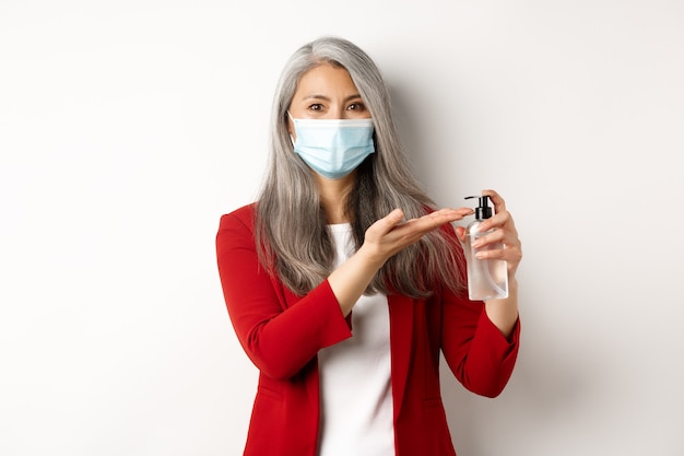 Concept de Covid, pandémie et entreprise. Gestionnaire féminine asiatique en masque médical à l'aide de désinfectant pour les mains et souriant à la caméra, debout avec un antiseptique sur fond blanc.