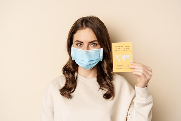 Concept de Covid et de personnes. Femme vaccinée portant un masque facial montrant un certificat de vaccination international covid, recommande de se faire vacciner, debout sur fond beige.