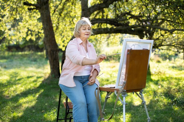 Concept de créativité, de passe-temps, d'art, de loisirs et de personnes. Portrait d'une artiste senior heureuse avec un pinceau, s'appuyant sur un chevalet à l'extérieur en plein air dans un parc verdoyant en été.