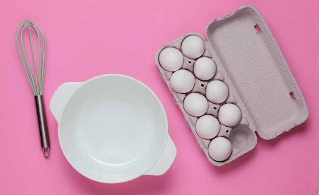 Concept de cuisine. Bol blanc vide avec fouet, plateau à oeufs sur fond de studio rose. Vue de dessus.