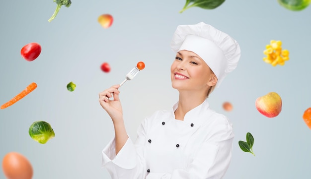 Photo concept de cuisine et de nourriture - chef féminin souriant, cuisinier ou boulanger avec fourchette et tomate sur des légumes en chute sur fond gris