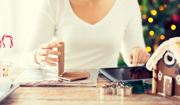 concept de cuisine, de personnes, de noël et de technologie - gros plan d'une femme souriante avec un ordinateur tablette faisant des maisons en pain d'épice à la maison