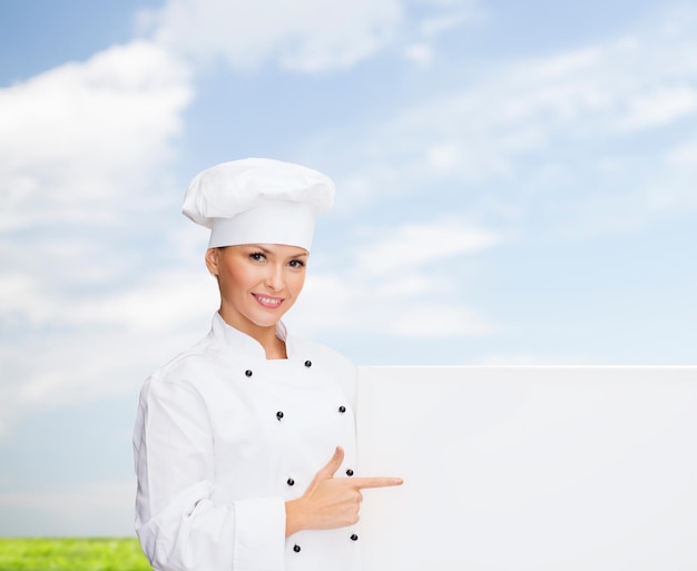 concept de cuisine, de publicité et de personnes - chef féminin souriant, cuisinier ou boulanger pointant le doigt vers un tableau blanc vierge sur fond de ciel bleu et d'herbe