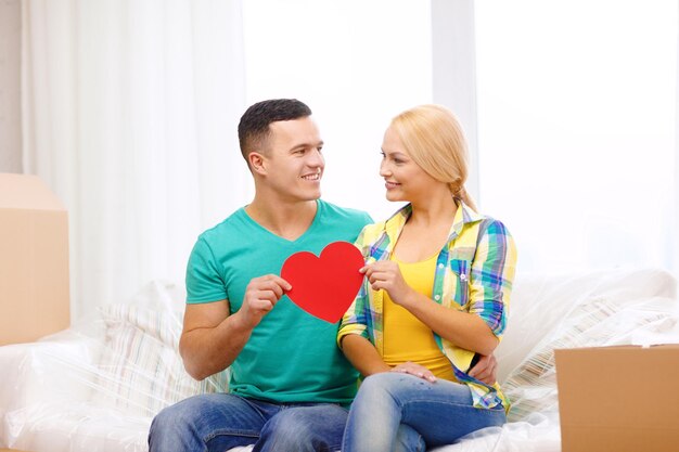concept de déménagement, de maison et de couple - couple souriant avec un coeur rouge sur un canapé dans une nouvelle maison