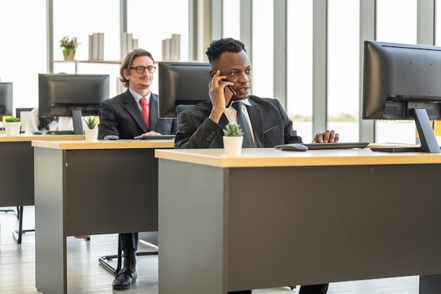 Concept De Diversité D'entreprise Et De Travail. Homme D'affaires Africain Parlant Sur Appel Avec Smartphone Mobile Et Travaillant Avec Un Ordinateur De Bureau.