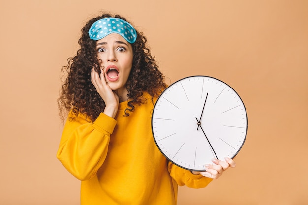 Concept du matin. Belle jeune femme frisée étonnée choquée posant sur fond beige avec horloge et masque de sommeil.