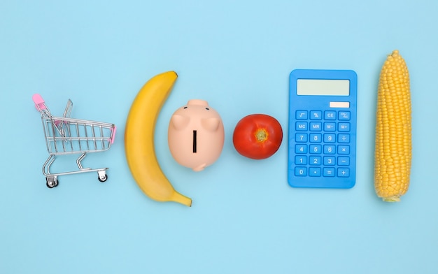 Le concept d'économiser sur la nourriture, faire du shopping. Chariot de supermarché, calculatrice, tirelire, légumes et fruits sur fond bleu. composition minimaliste