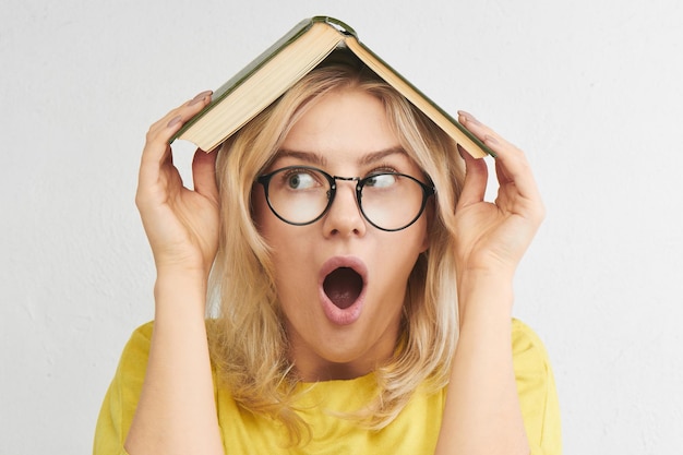 Concept éducatif. Une étudiante européenne à lunettes tient un manuel sur la tête avec une bouche ouverte surprise, le stress des études. Fond de studio blanc