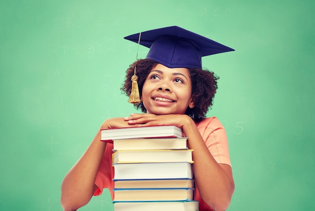 concept d'éducation, d'école, de connaissances et de personnes - heureuse étudiante afro-américaine souriante en chapeau de célibataire avec des livres assis à table et rêvant sur fond de tableau de craie verte