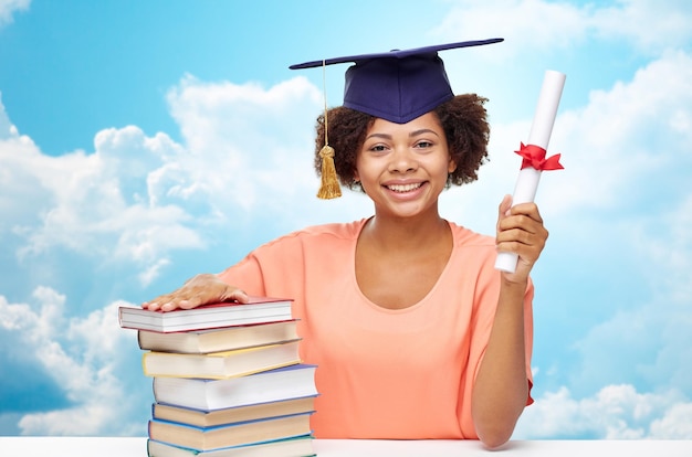 Concept d'éducation, d'école, de connaissances, de remise des diplômes et de personnes - heureuse étudiante afro-américaine souriante en bonnet de célibataire avec livres et diplôme assis à table sur fond bleu ciel et nuages