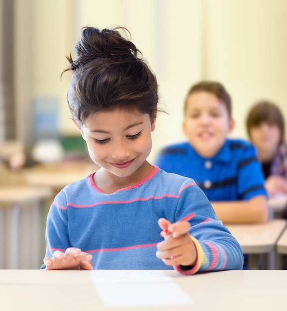 concept d'éducation, d'école primaire et d'enfants - heureuse petite fille étudiante avec stylo et papier écrivant sur fond de classe et de camarades de classe