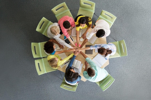 concept d'éducation, d'école, de travail d'équipe et de personnes - groupe d'étudiants internationaux avec des livres et des ordinateurs tablettes mettant les mains les unes sur les autres assis à table