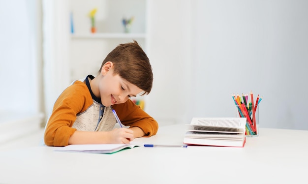 concept d'éducation, d'enfance, de personnes, de devoirs et d'école - garçon étudiant souriant avec un livre écrit sur un cahier à la maison