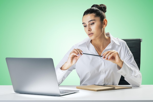 Concept d'éducation en ligne et de formation Internet avec une jolie jeune femme assise à une table blanche regardant un écran d'ordinateur portable et portant un crayon dans les mains sur fond de mur vert clair abstrait