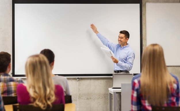 concept d'éducation, de lycée, de technologie et de personnes - enseignant souriant avec bloc-notes, ordinateur portable debout devant les élèves et montrant quelque chose sur un tableau blanc dans la salle de classe