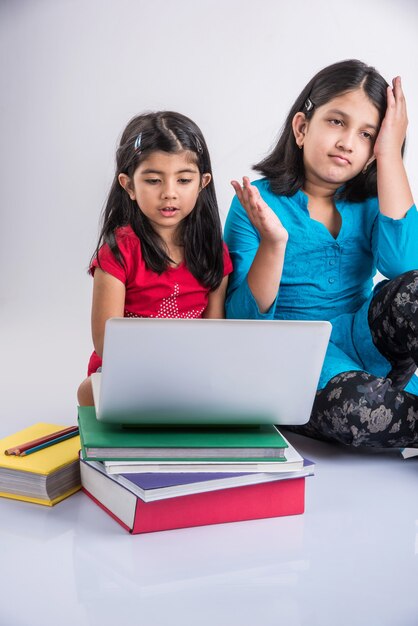 Concept d'éducation à la maison - 2 jolies petites filles indiennes ou asiatiques ludiques qui étudient ou font leurs devoirs sur un ordinateur portable sur une pile de livres tout en étant assises par terre à la maison. Isolé sur fond blanc