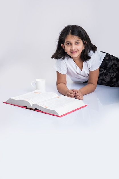 Concept d'éducation à la maison - Jolie petite fille indienne ou asiatique étudiant avec une pile de livres et une tasse à café assise par terre à la maison. Isolé sur fond blanc