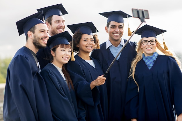 concept d'éducation, de remise des diplômes, de technologie et de personnes - groupe d'étudiants internationaux heureux dans des planches de mortier et des robes de célibataire prenant un selfie par smartphone à l'extérieur