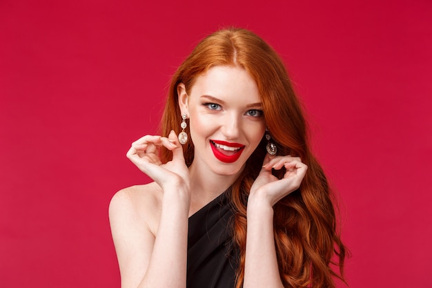 Concept d'élégance, de beauté et de mode. Gros plan, portrait, de, impertinent, beau, séduisant, jeune, rousse, femme, toucher, elle, boucles d'oreilles, sûr de soi, flirter, sur, mur rouge