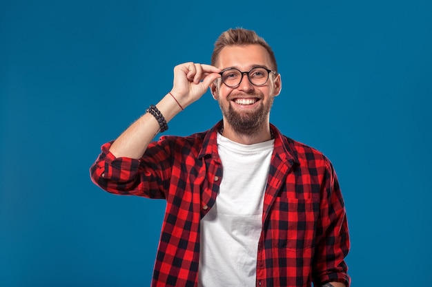 Concept émotionnel et humain : jeune homme barbu en chemise à carreaux. Style hipster. Tourné en studio sur fond bleu. Copiez l'espace. Le gars ajuste ses lunettes