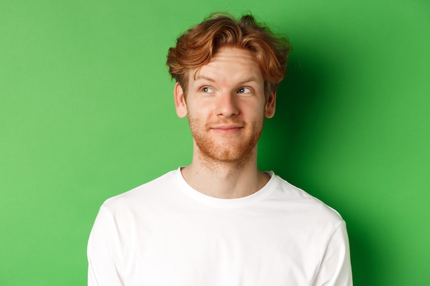 Concept d'émotions et de mode. Gros plan du modèle masculin beau rousse avec barbe et coupe de cheveux en désordre, regardant à gauche et souriant, fond vert.