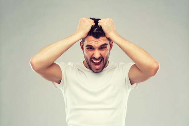 Concept d'émotions, de stress, de folie et de personnes - homme criant fou déchirant ses cheveux en t-shirt sur fond gris