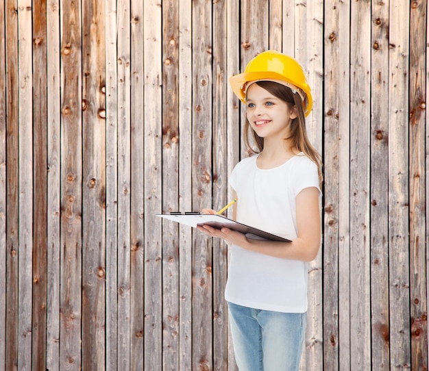 concept d'enfance, de construction, d'architecture, de bâtiment et de personnes - petite fille souriante dans un casque de protection avec presse-papiers prenant des notes