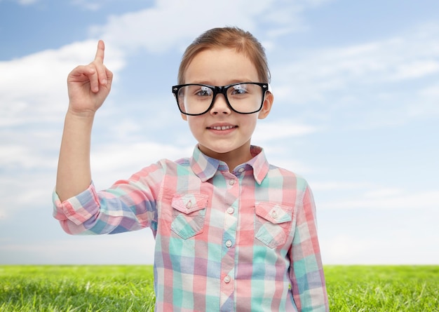 concept d'enfance, d'école, d'éducation, de vision et de personnes - heureuse petite fille à lunettes pointant le doigt vers le haut sur fond de ciel bleu et de champ vert